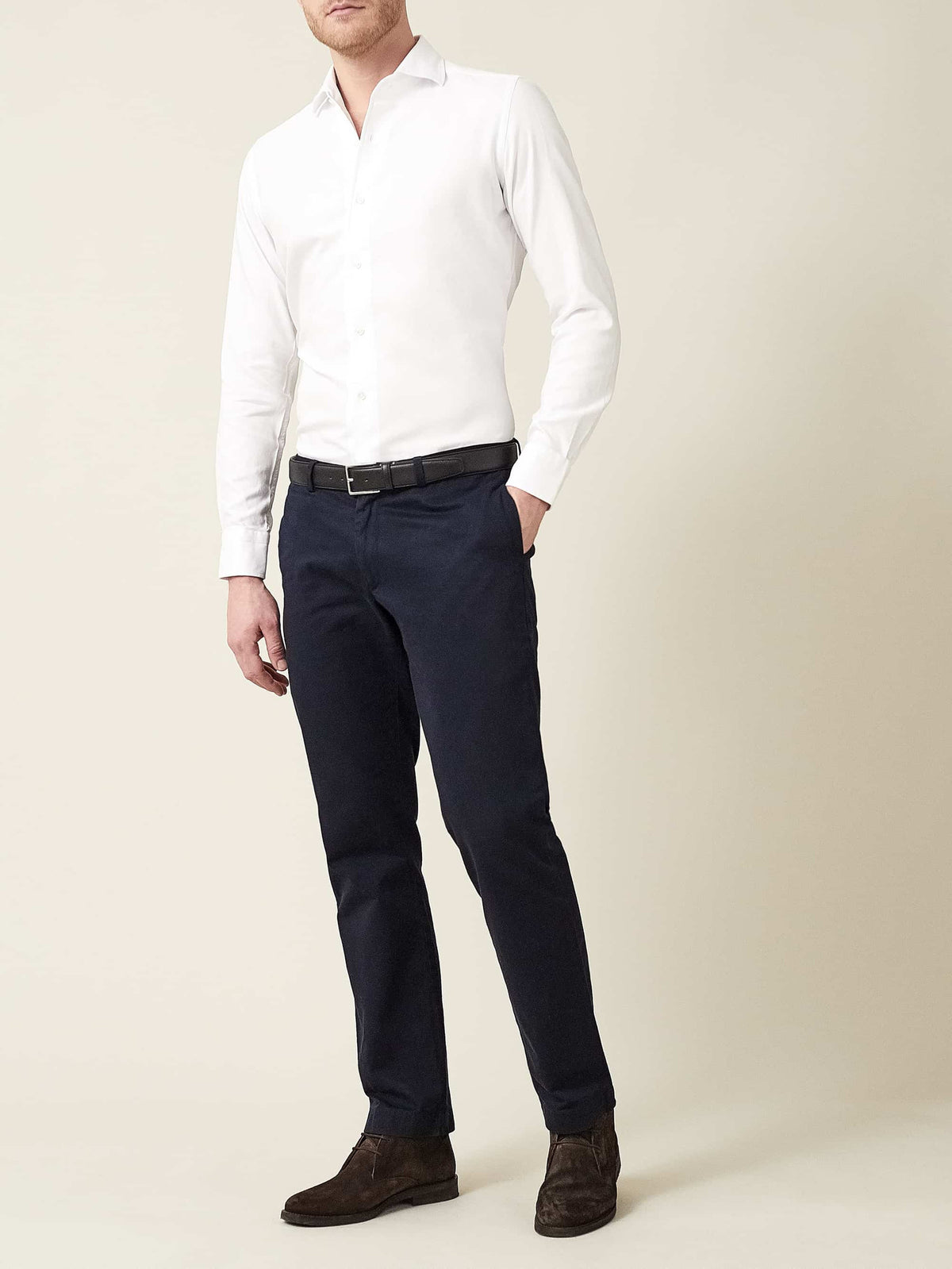A model wears the White Oxford Cotton Shirt, tucked into navy trousers and styled with a black belt and brown suede shoes.