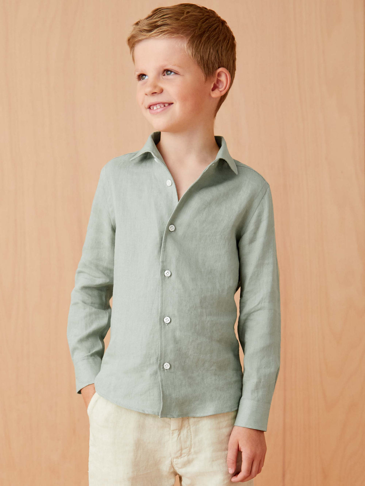 A young boy wearing the Portofino Junior Linen Shirt in Sage Green, shown from the front against a light wood background.