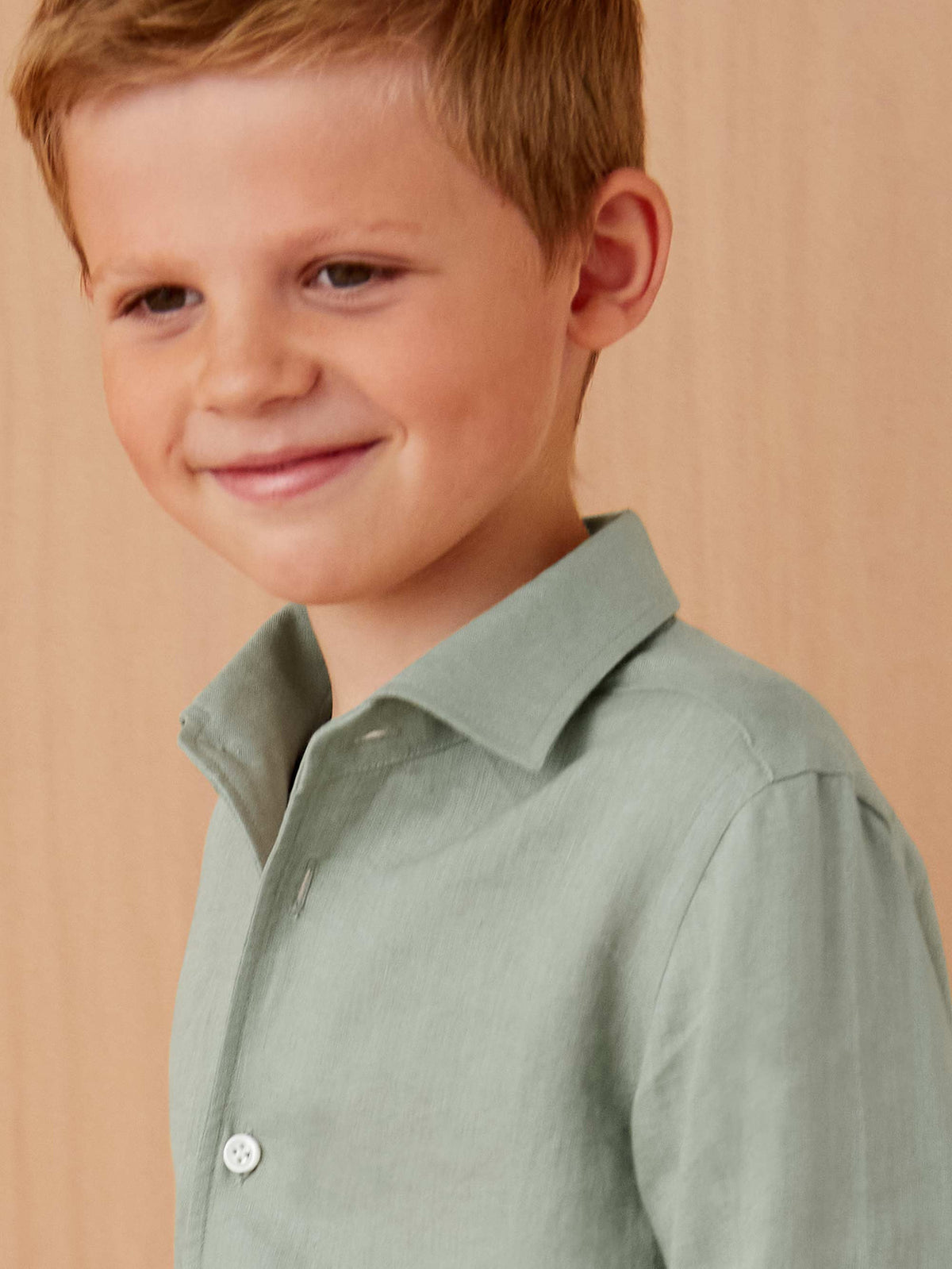 A close-up of a smiling young boy wearing the sage green Portofino Junior Linen Shirt.