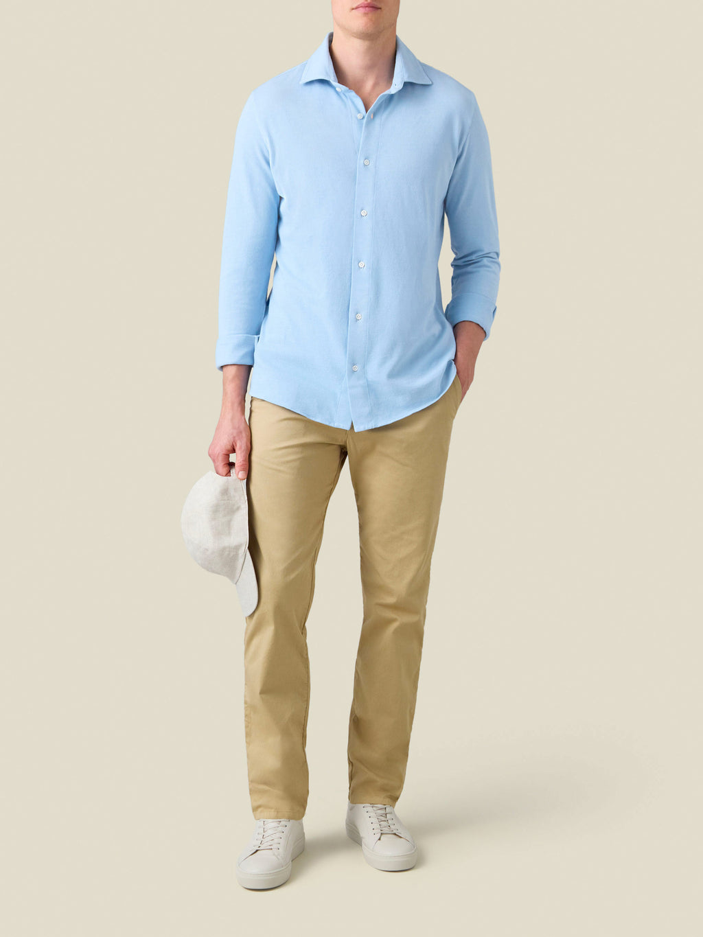Man wears the light blue Siena Piqué Shirt with tan chinos and white sneakers, holding a white cap.