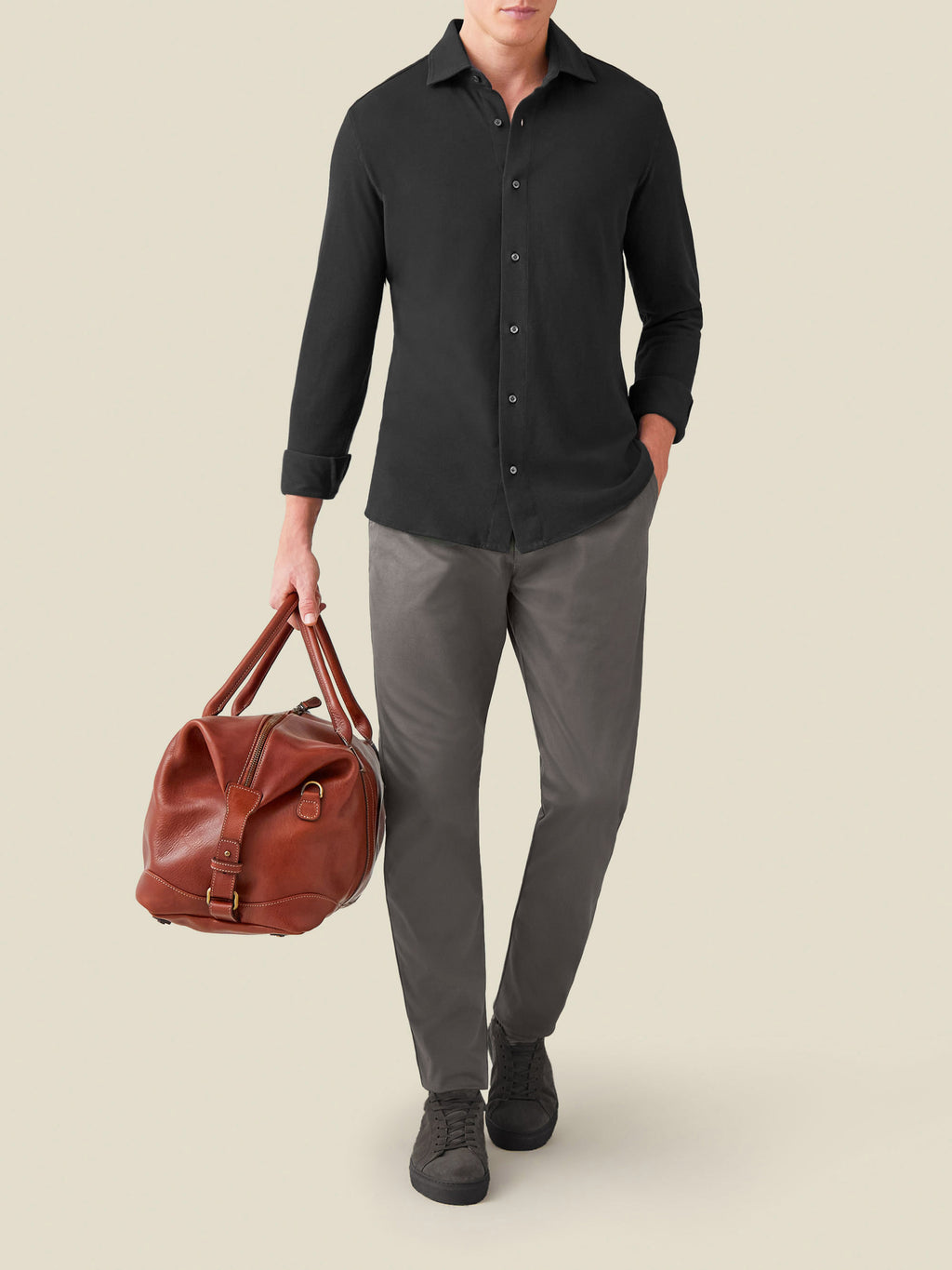 Model wearing the Siena Piqué Shirt in Charcoal Grey, paired with grey trousers and carrying a brown leather travel bag.