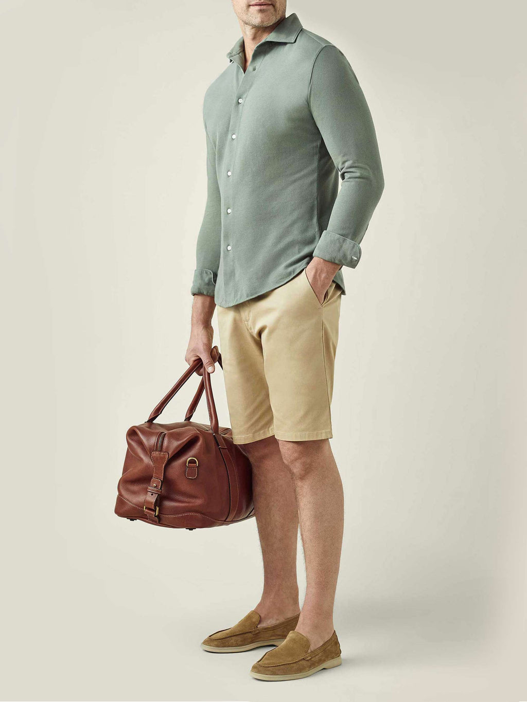 Model wears the Siena Piqué Shirt in sage green, styled with beige shorts and holding a brown leather travel bag.