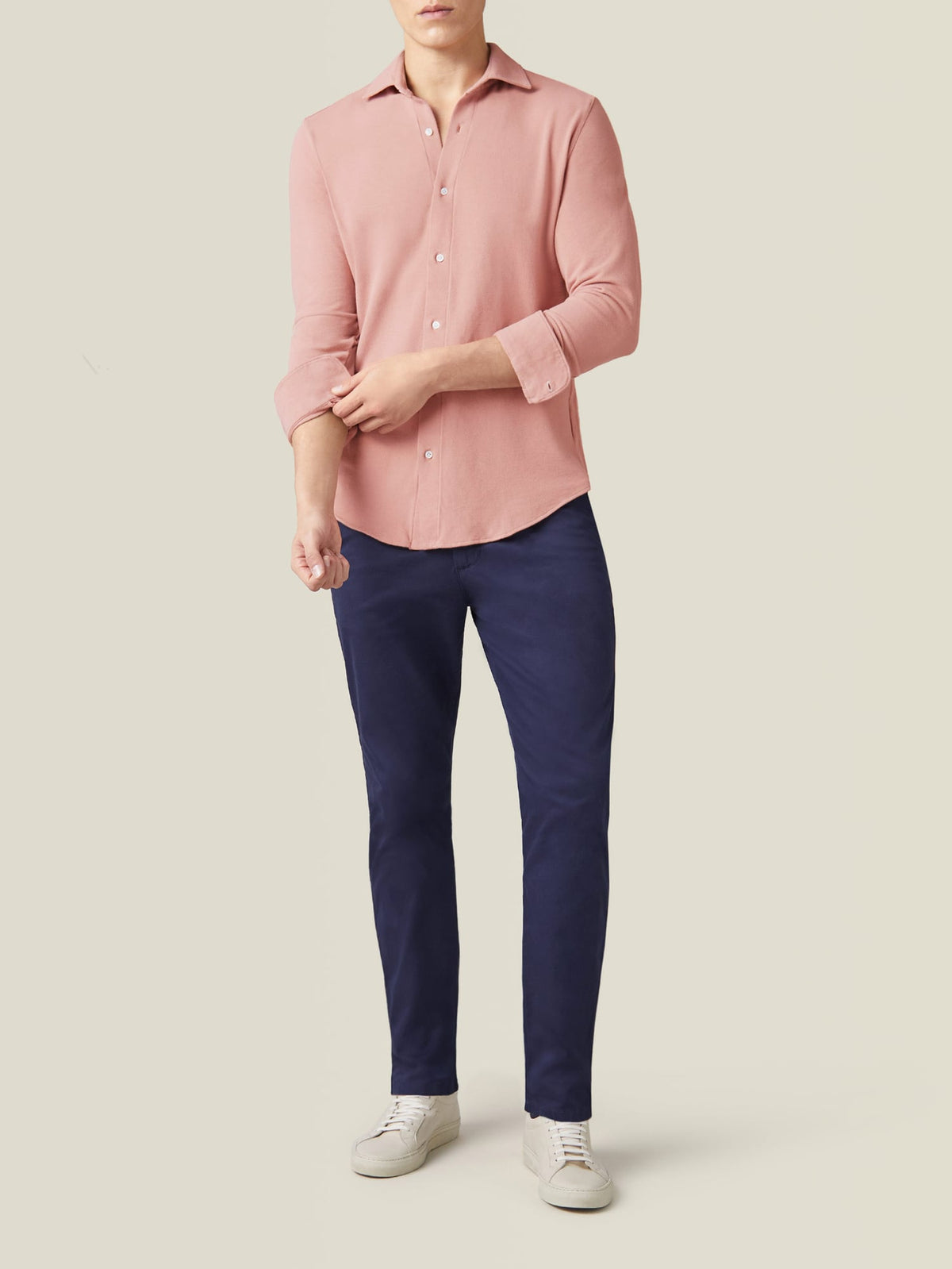 A model stands facing forward wearing the Siena Piqué Shirt in Peach, paired with navy trousers and white trainers.