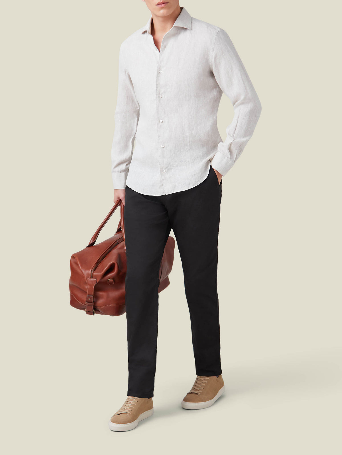Man wearing black lightweight cotton chinos with a light grey shirt and tan leather sneakers, holding a brown weekender bag.