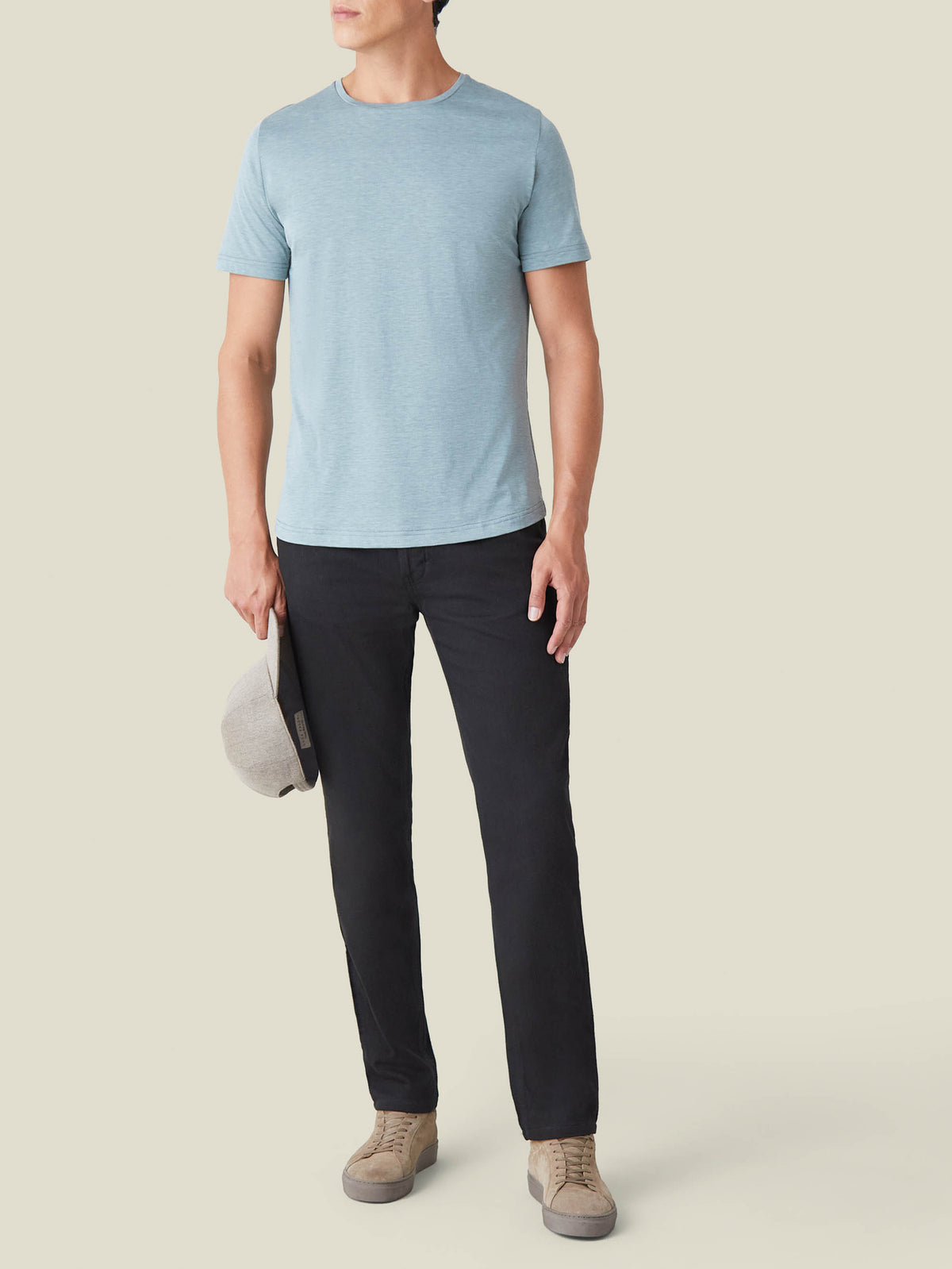 Model wears Luca Faloni black tapered fit jeans with a light blue t-shirt and beige suede shoes, holding a grey cap.