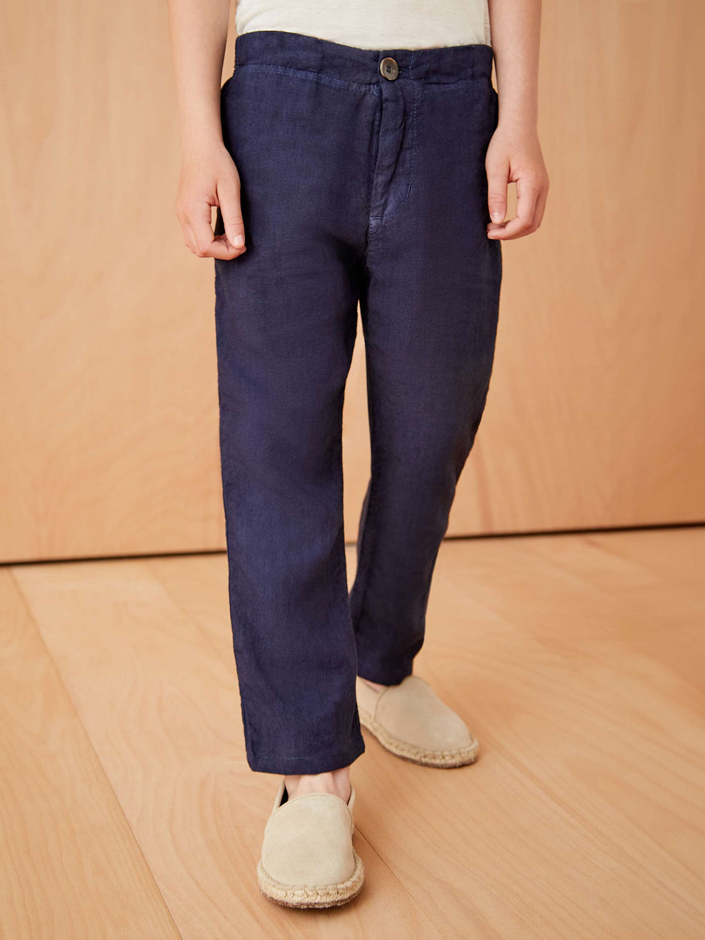 Front view of a boy wearing the navy blue Lipari Junior Linen Trousers with beige espadrilles against a wooden backdrop.
