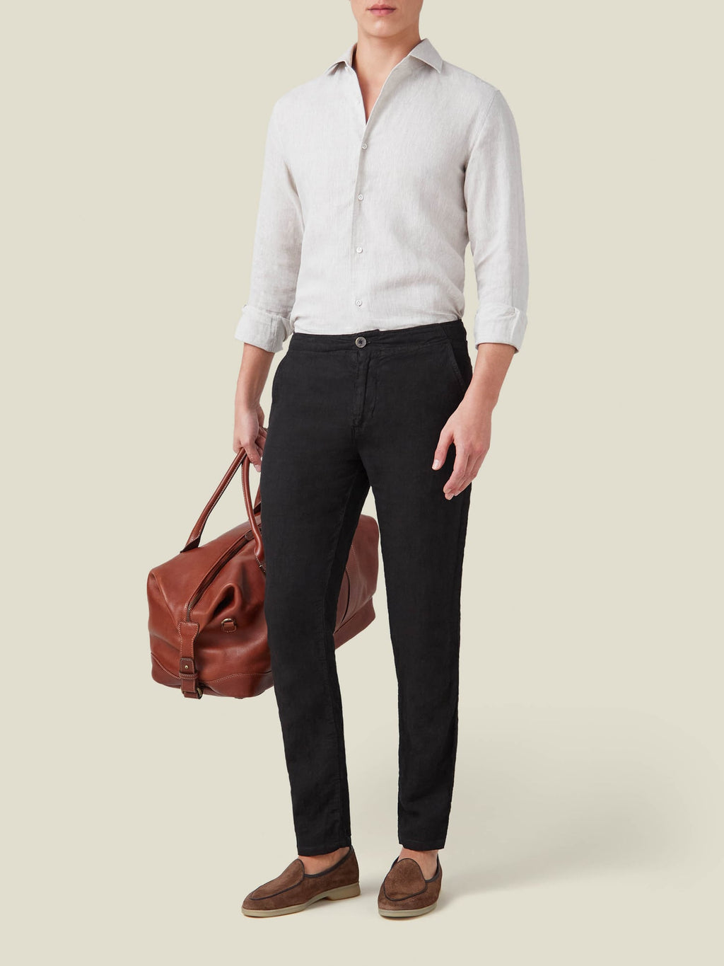 A man styles the black Lipari Linen Trousers with a light linen shirt, brown loafers, and a leather weekender bag.