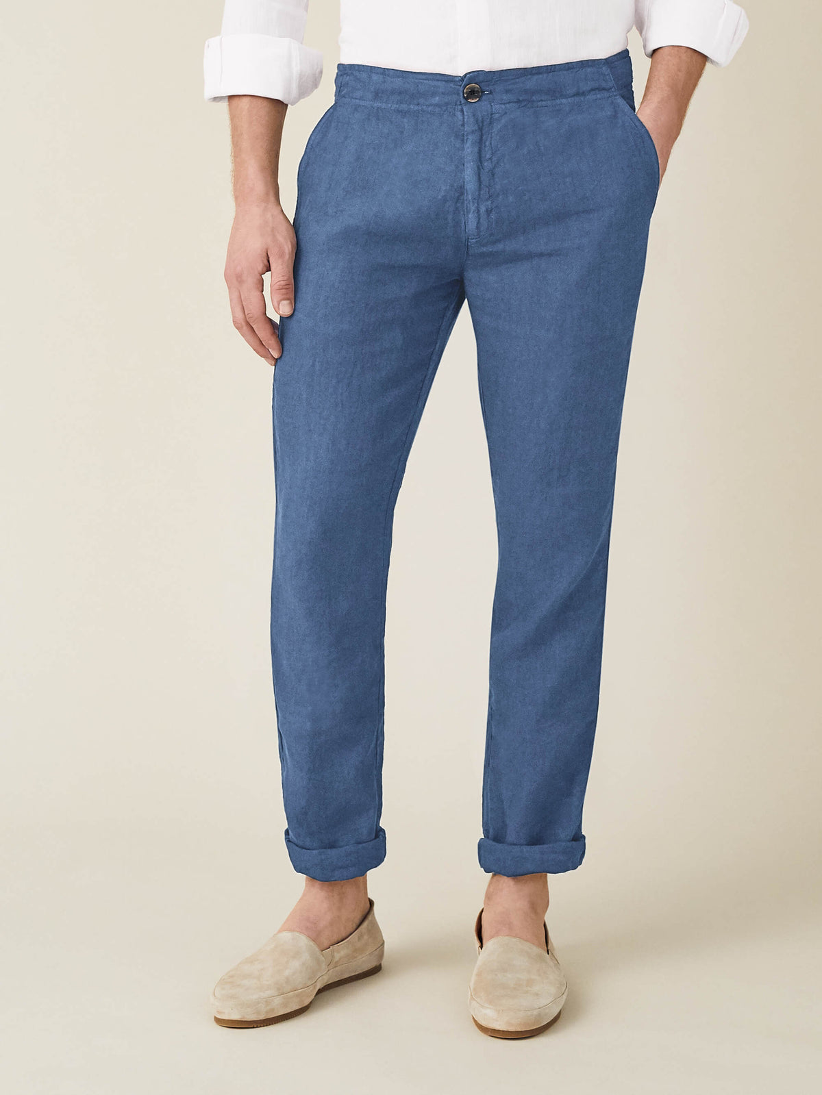 A man wears the Lipari Linen Trousers in Capri Blue with rolled cuffs and beige loafers, shown from the front.
