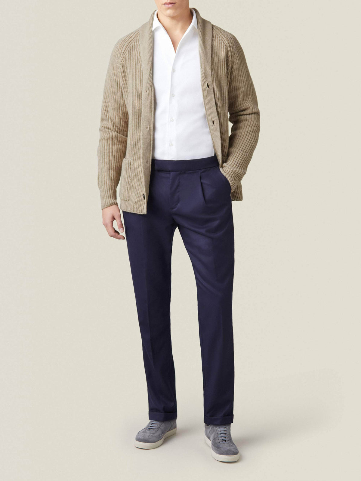 A man models Luca Faloni's Wool Pleated Trousers in midnight blue, paired with a white shirt and a beige cardigan.