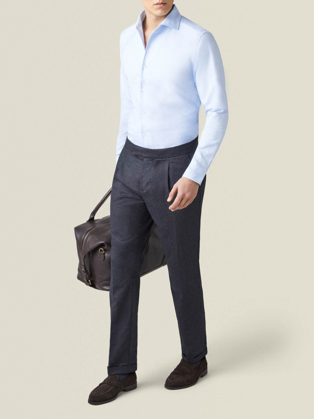 A man wearing Luca Faloni's charcoal grey wool pleated trousers with a light blue shirt, holding a dark brown leather bag.