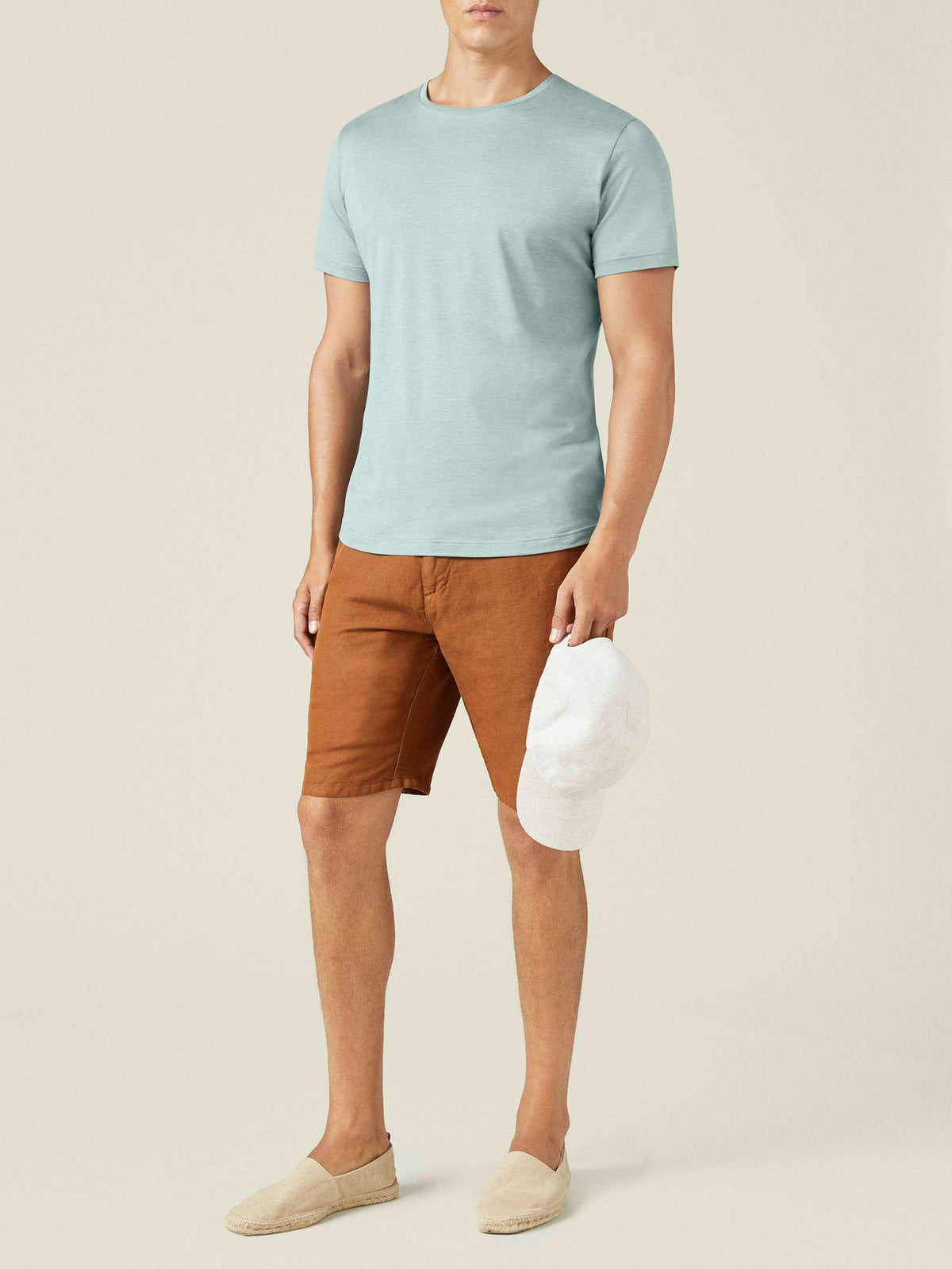 Full-length view of a model in a marine green silk-cotton t-shirt, brown shorts, and espadrilles, holding a white cap.
