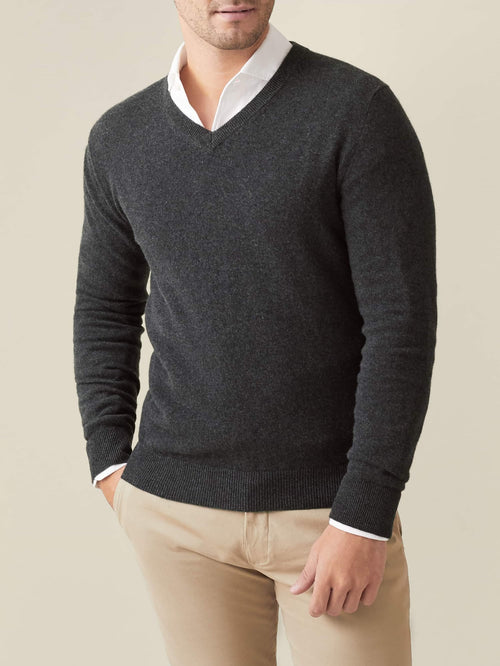 A man models the Luca Faloni charcoal grey cashmere v-neck jumper, layered over a white collared shirt and beige trousers.