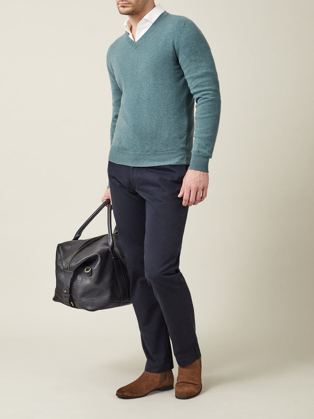 Man wears a marine green cashmere V neck sweater over a white shirt with navy trousers, brown boots, holding a black bag.