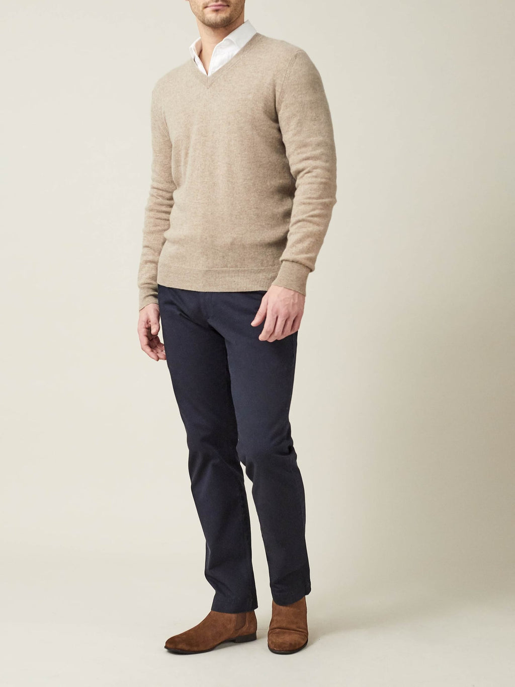Man wears a camel beige cashmere V-neck sweater, layered over a white shirt, paired with navy trousers and brown suede boots.