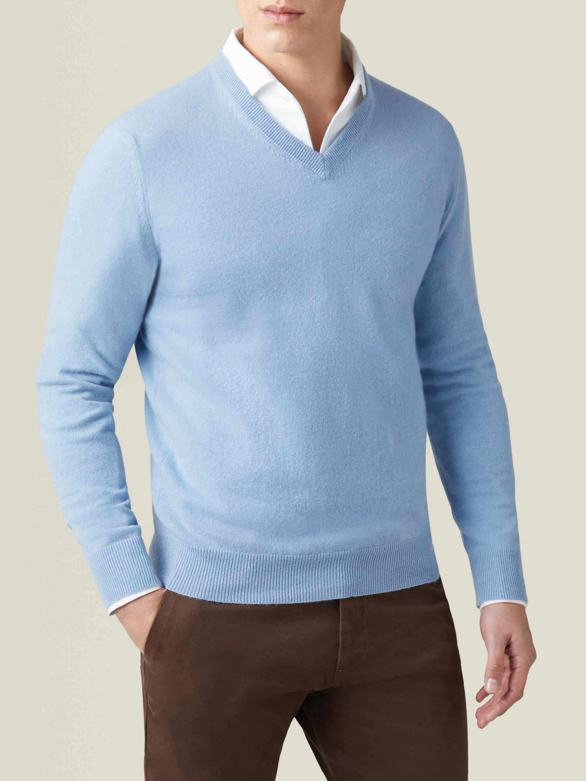 Man wearing a sky blue pure cashmere V-neck sweater, layered over a white shirt and paired with brown trousers.