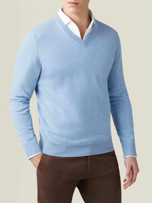 Man wearing a sky blue pure cashmere V-neck sweater, layered over a white shirt and paired with brown trousers.
