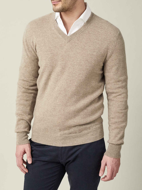 Model wearing a camel beige cashmere V-neck jumper, styled over a white collared shirt and dark trousers.