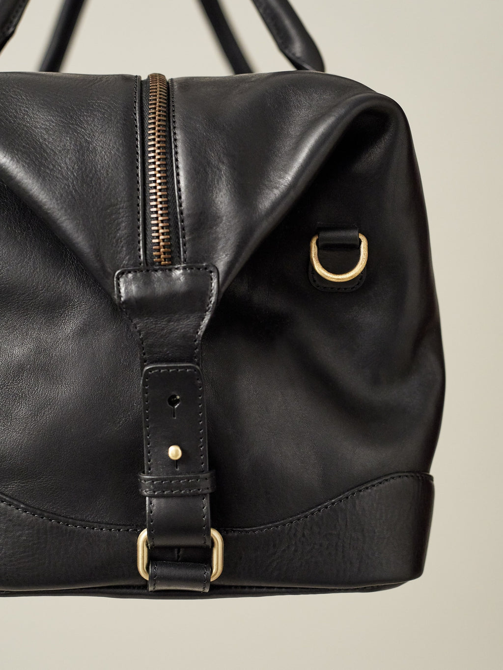 Detail of the Luca Faloni Weekender bag in black leather, showing the brass zipper, side buckle, and shoulder strap D-ring.