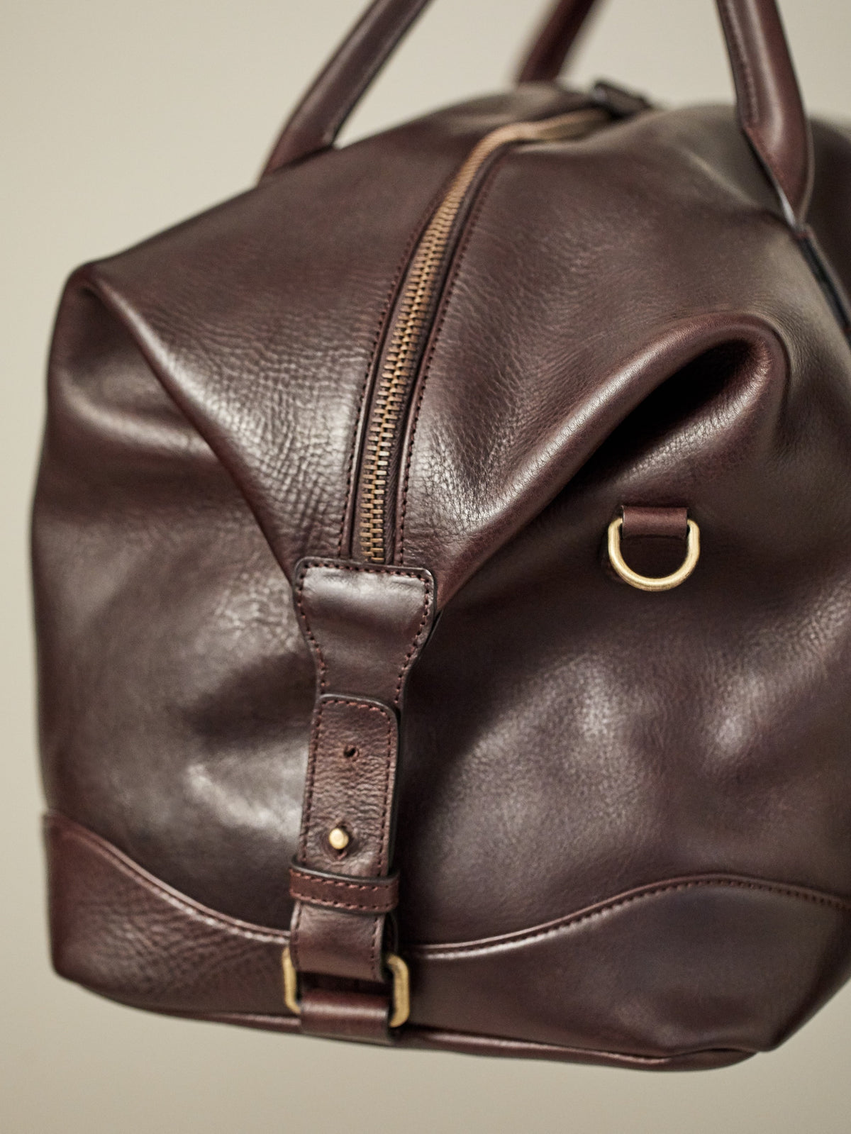 A close-up shot of the Luca Faloni Weekender bag in chocolate brown leather, detailing the brass zipper and side strap.