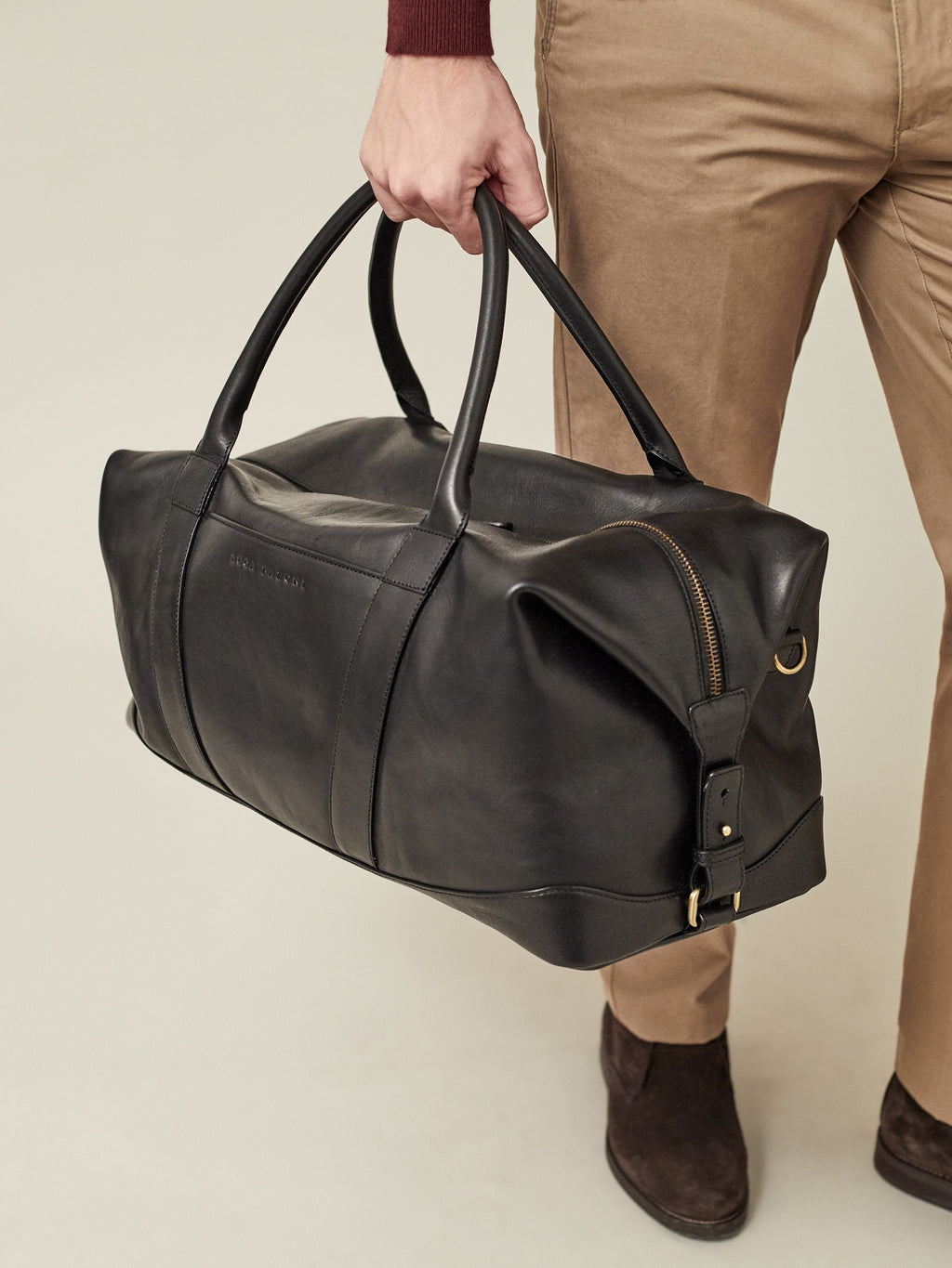A man holding the Luca Faloni Weekender bag in black leather, styled with tan trousers and dark brown suede shoes.