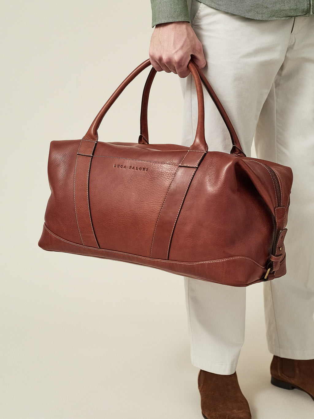 A close-up of a man holding the Luca Faloni Weekender bag in Caramel Brown leather by its top handles.