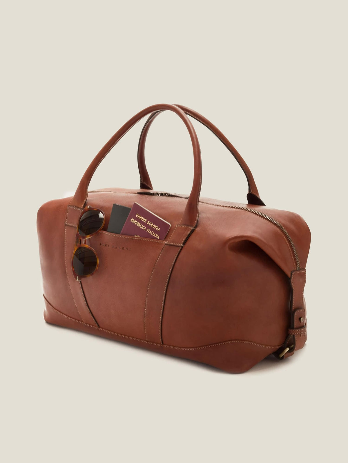 The Luca Faloni Weekender bag in caramel brown leather, shown with sunglasses and a passport in the external sleeve pocket.