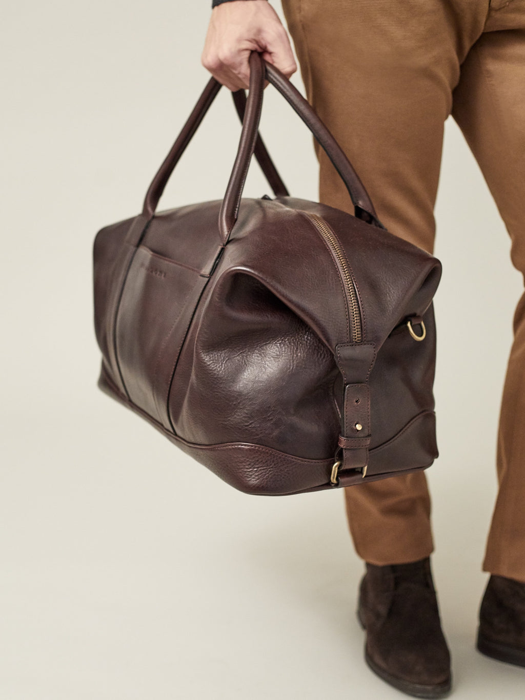 Model holding the Luca Faloni Weekender bag in chocolate brown leather, showing a side view of the travel bag.