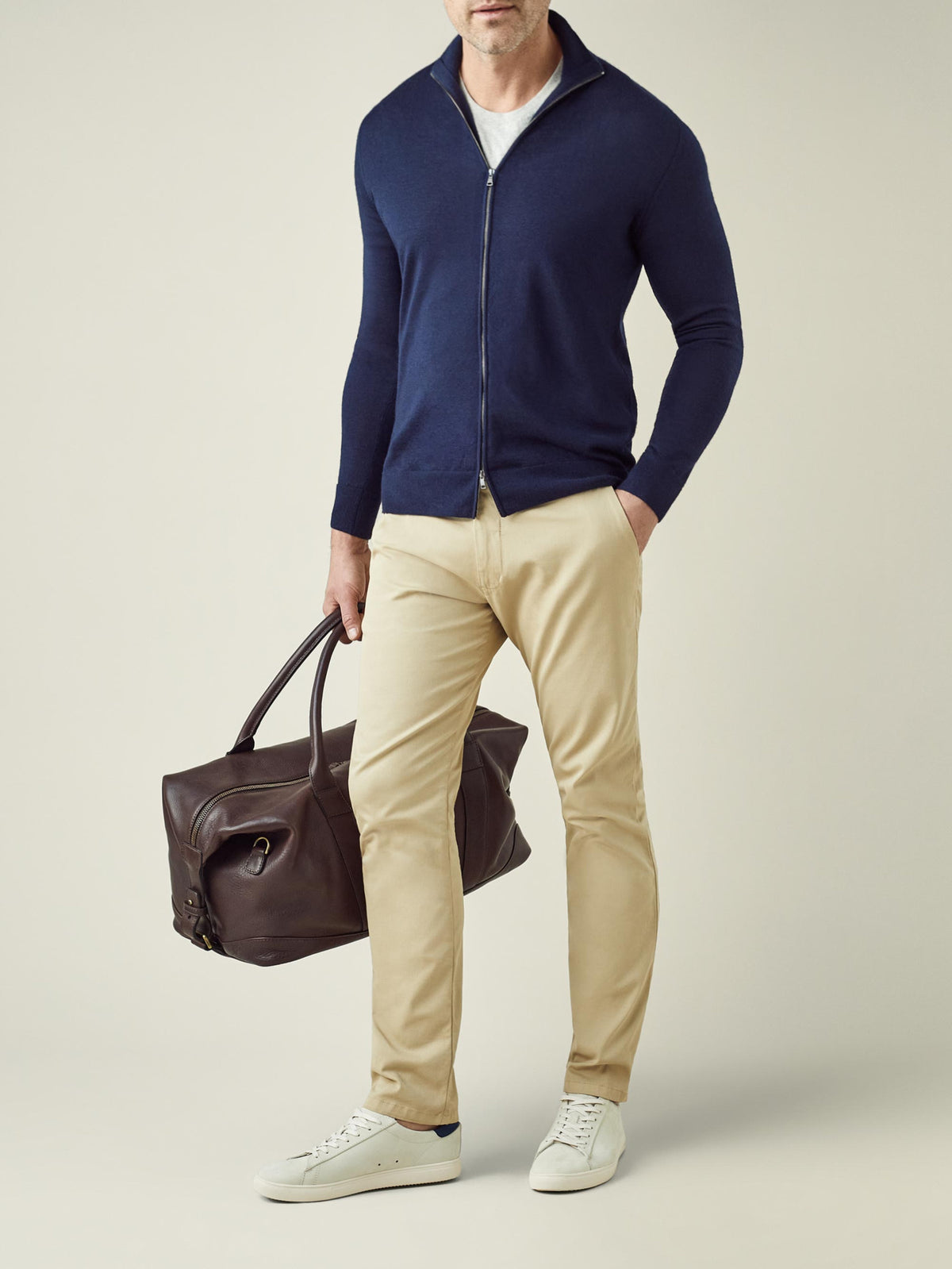 A man models the Luca Faloni navy blue silk-cashmere zip cardigan, styled with beige chinos and holding a travel bag.