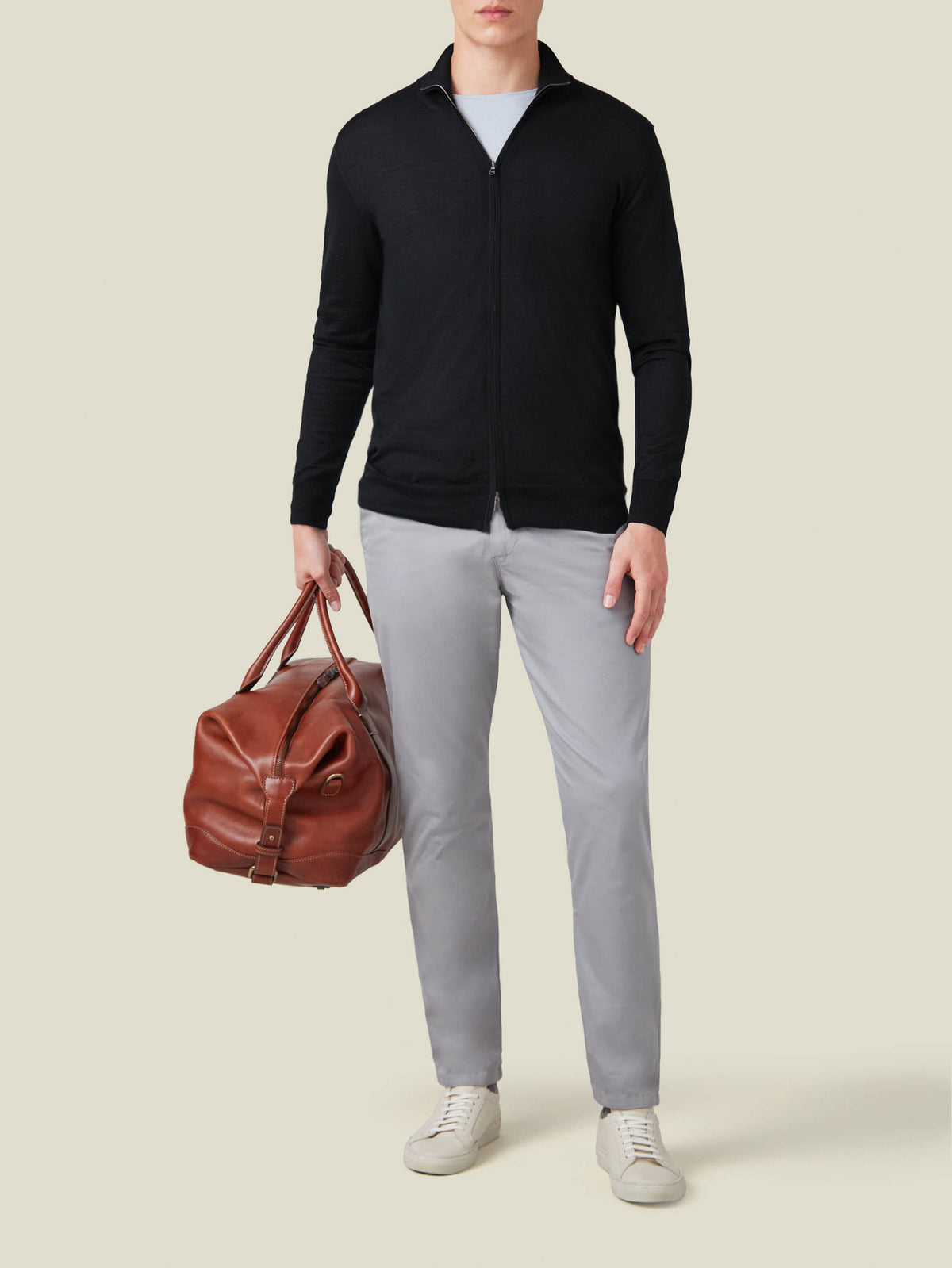 A model wearing the Luca Faloni Black Silk-Cashmere Zip Cardigan, styled with grey trousers and a brown leather travel bag.