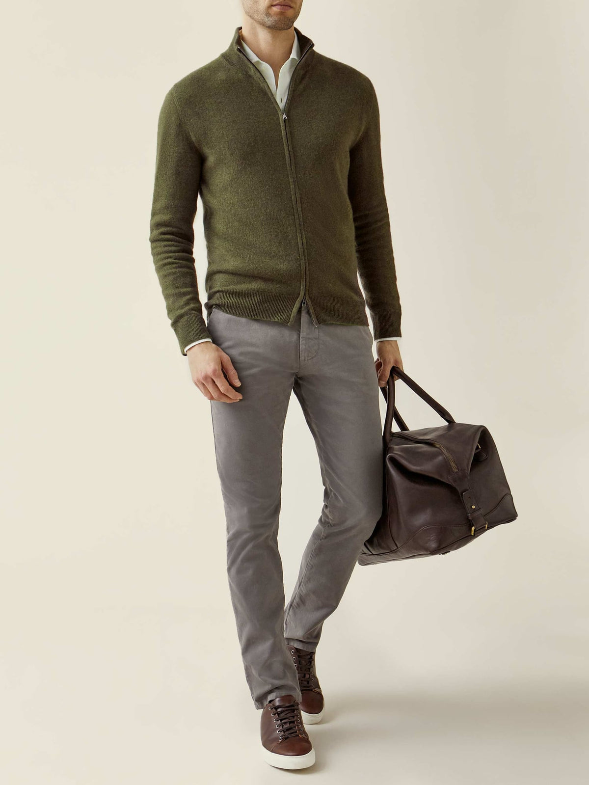 Model wears a Hunting Green Cashmere Zip Cardigan with grey trousers, brown sneakers, and a leather travel bag.