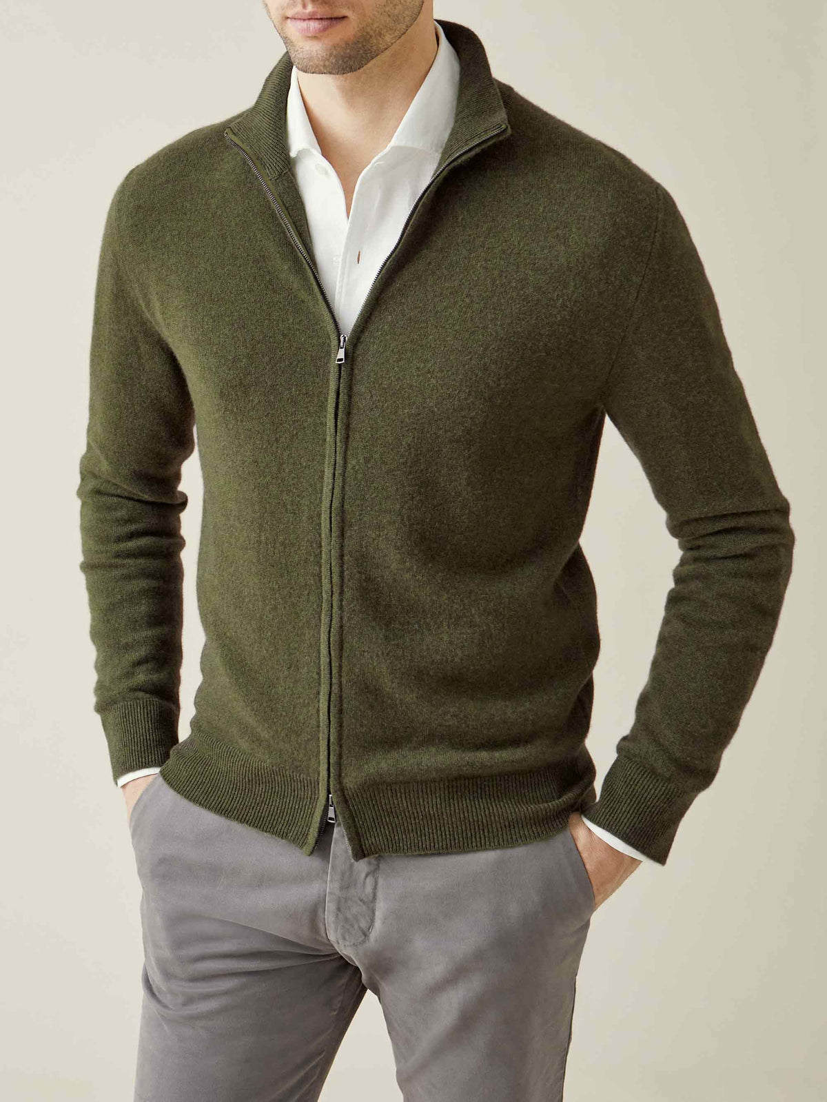 A man models the Luca Faloni Cashmere Zip Cardigan in Hunting Green, over a white shirt and paired with grey trousers.