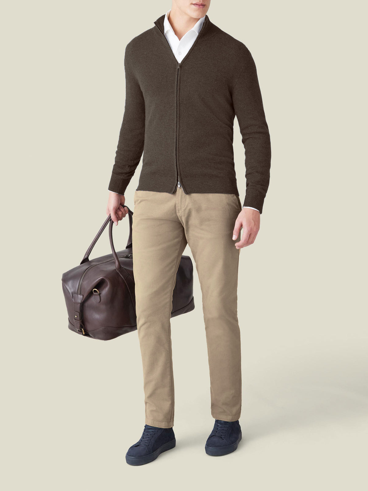 Man wearing a Chocolate Brown Cashmere Zip Cardigan with beige trousers, holding a brown leather travel bag.