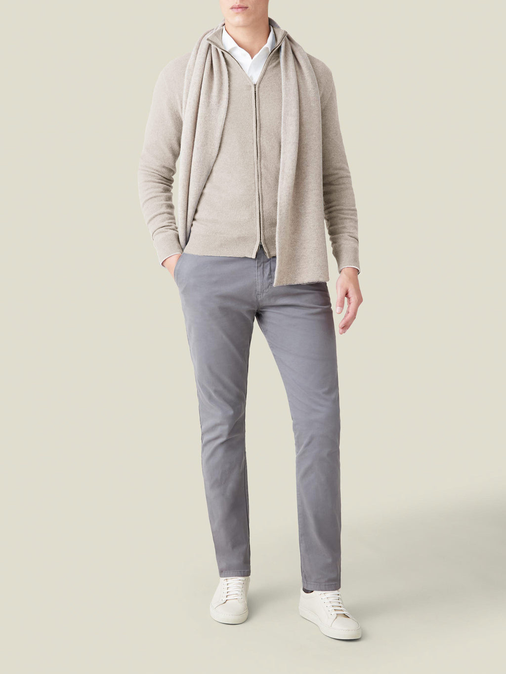 A model wears the Luca Faloni Almond Cashmere Zip Cardigan styled with a matching scarf, grey trousers, and white sneakers.