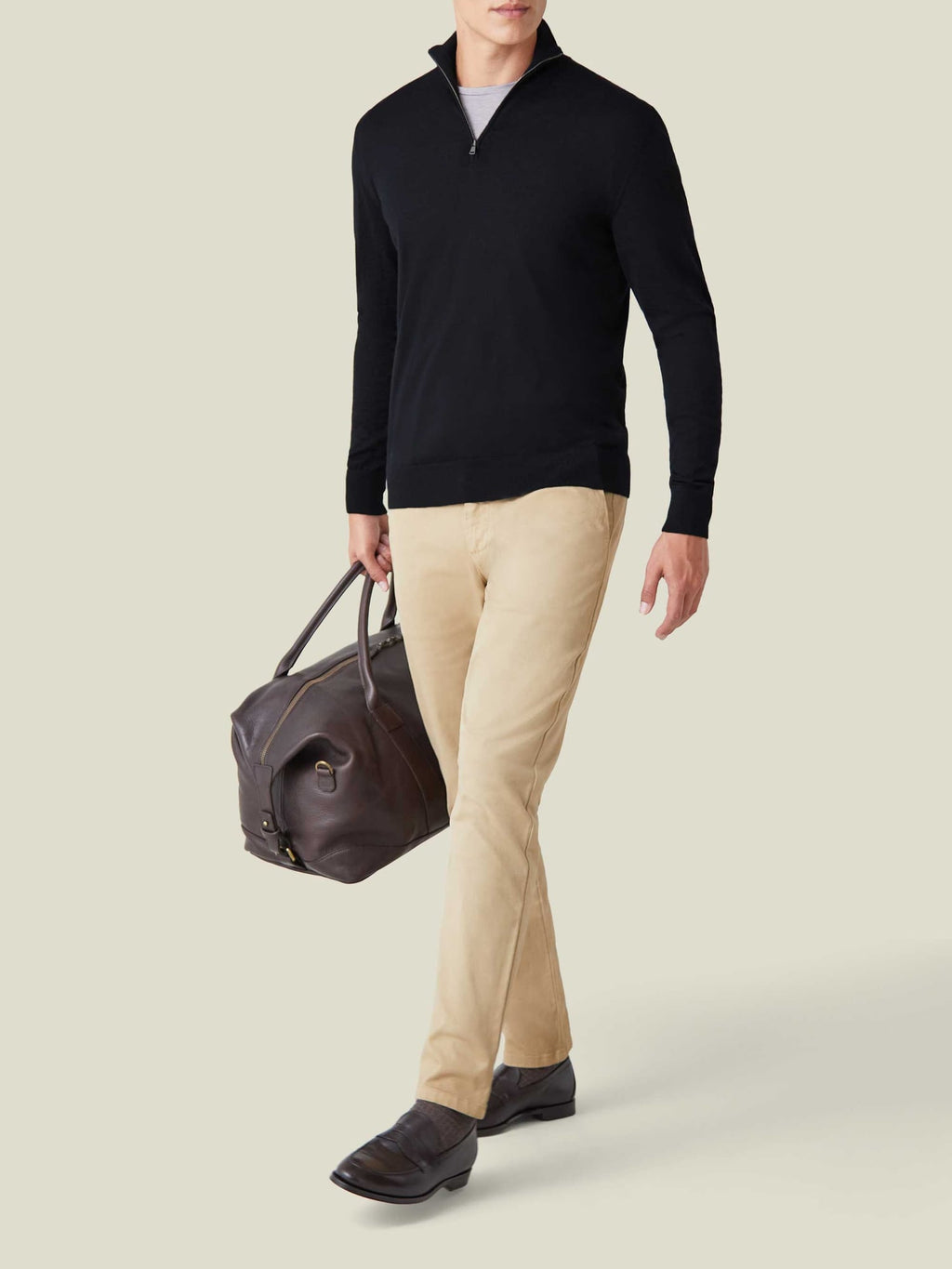 Model wearing the black silk-cashmere quarter-zip with beige chinos, holding a leather travel bag.