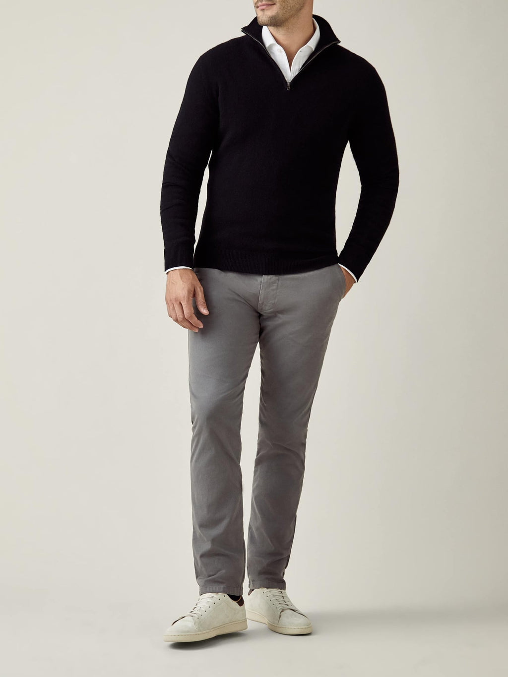 A man models a black cashmere zip-up sweater over a white collared shirt, paired with grey trousers and white sneakers.