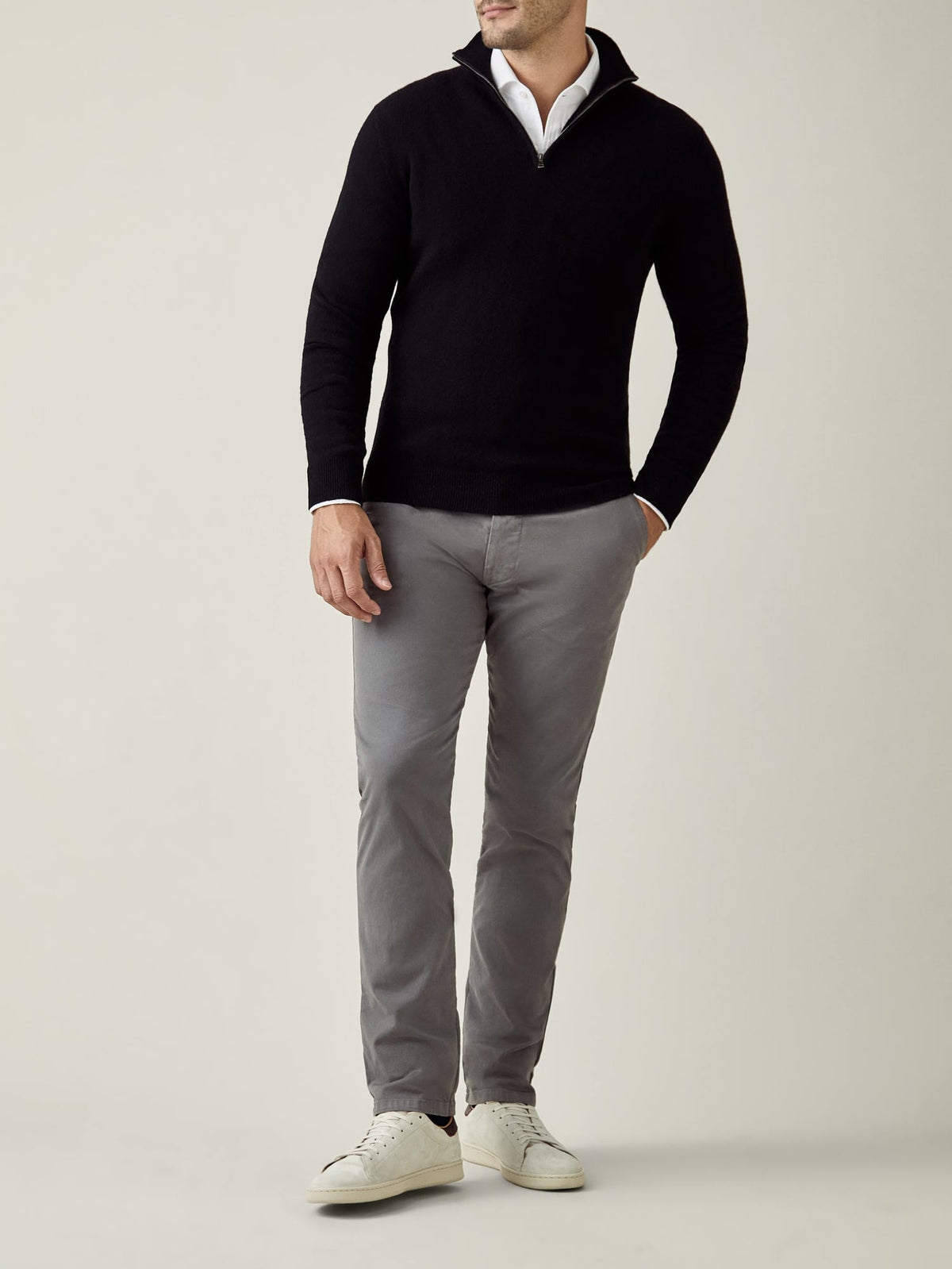 A man models a black cashmere zip-up sweater over a white collared shirt, paired with grey trousers and white sneakers.