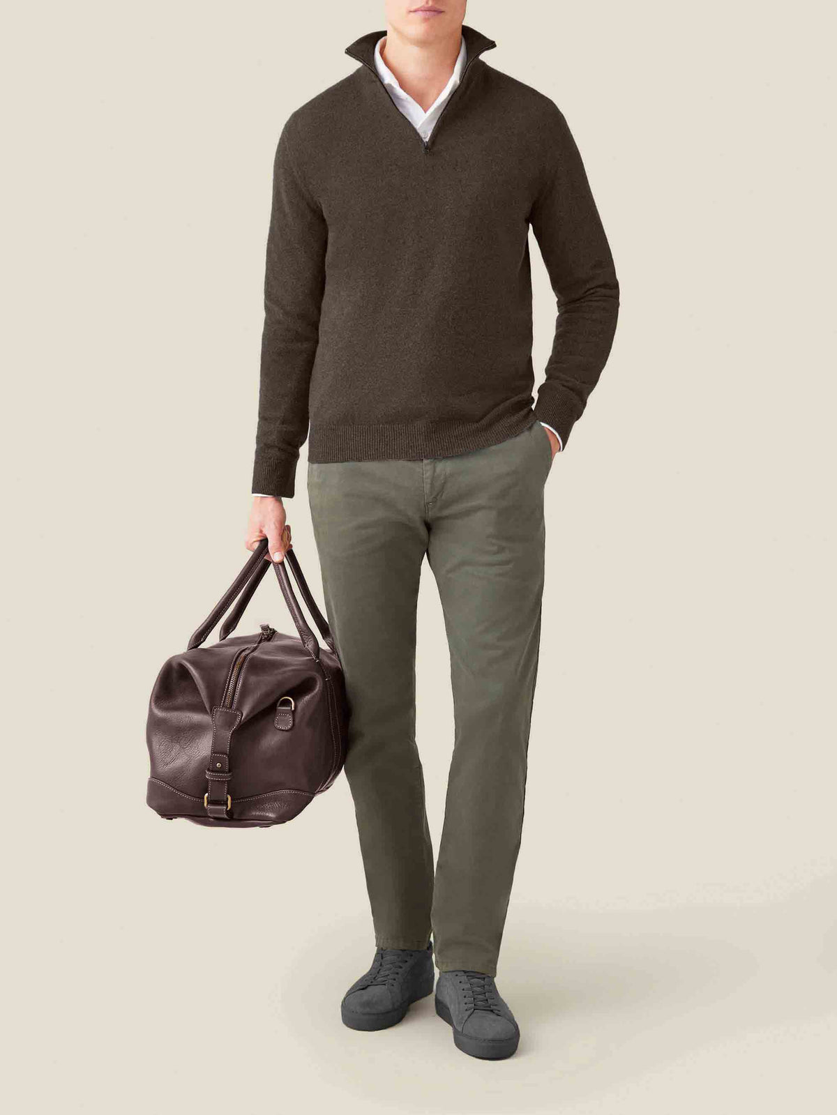 Model wears the Luca Faloni chocolate brown cashmere zip-up layered over a shirt with green trousers and a leather holdall.