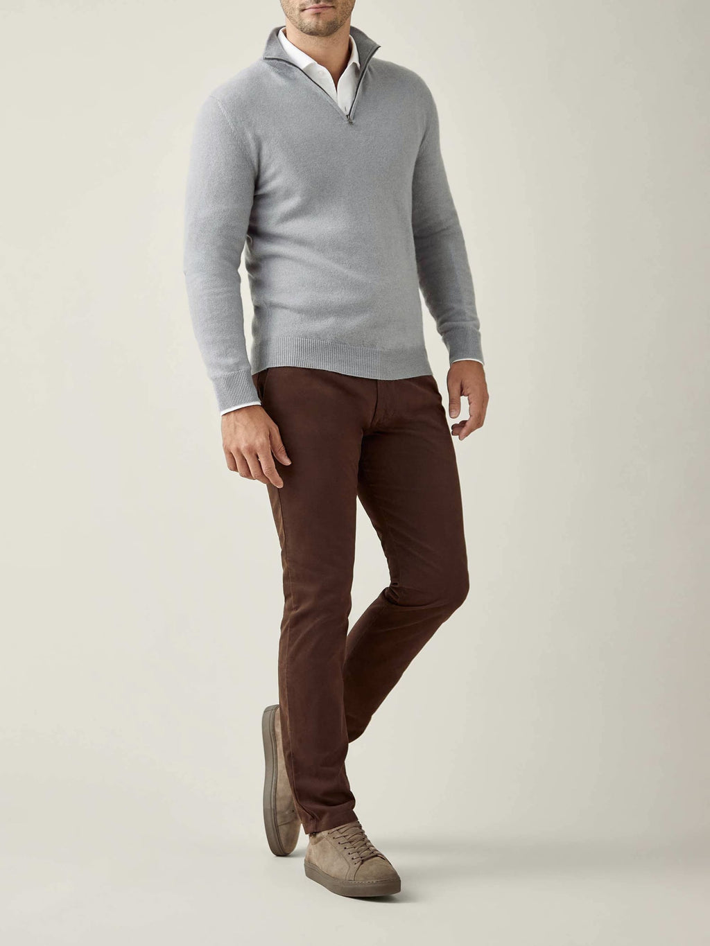 A man models the misty grey cashmere zip-up, worn over a white collared shirt with brown trousers and suede shoes.