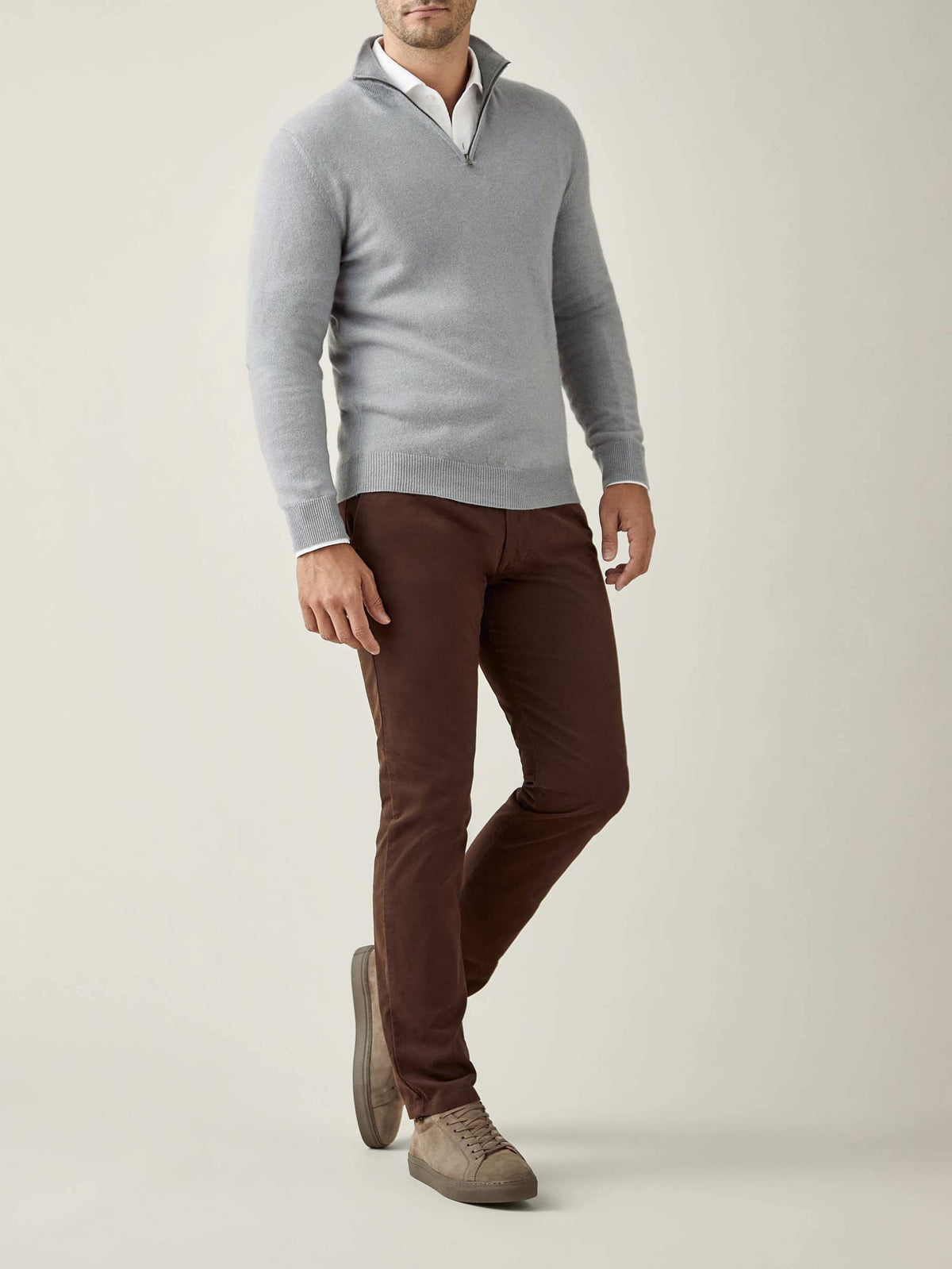 A man models the misty grey cashmere zip-up, worn over a white collared shirt with brown trousers and suede shoes.