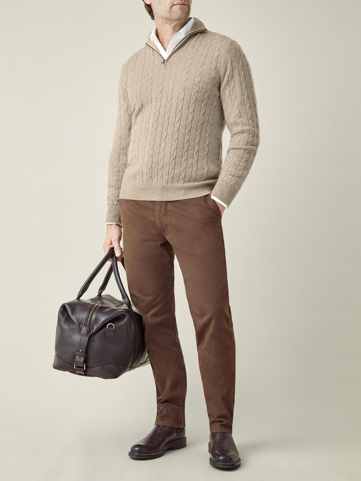 Man wearing the Luca Faloni Cable Knit Zip-up in camel beige with brown trousers and holding a leather duffle bag.