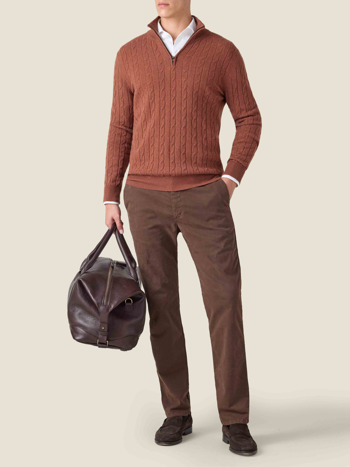 Model wears the rust Cable Knit Zip-up over a white shirt, with brown trousers, holding a dark brown leather weekend bag.