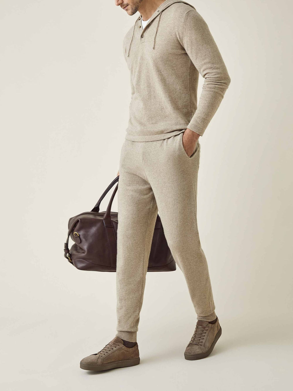 A model wears the Luca Faloni camel beige cashmere joggers and hoodie, styled for travel with a leather bag and sneakers.