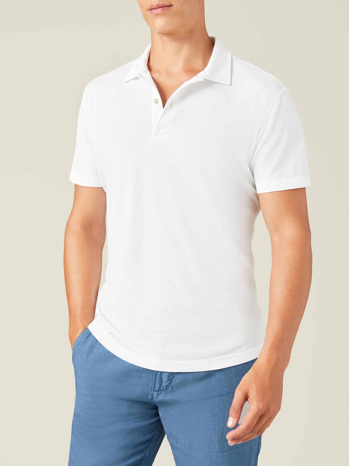 A man models the Luca Faloni white short-sleeved Classic Piqué Polo with his hand in the pocket of his blue trousers.