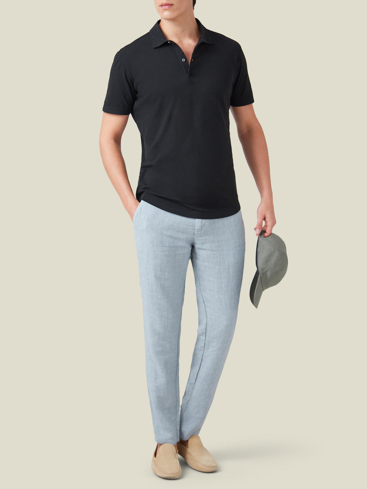 Front view of a model wearing the black Classic Piqué Polo with light blue trousers and beige loafers, holding a cap.