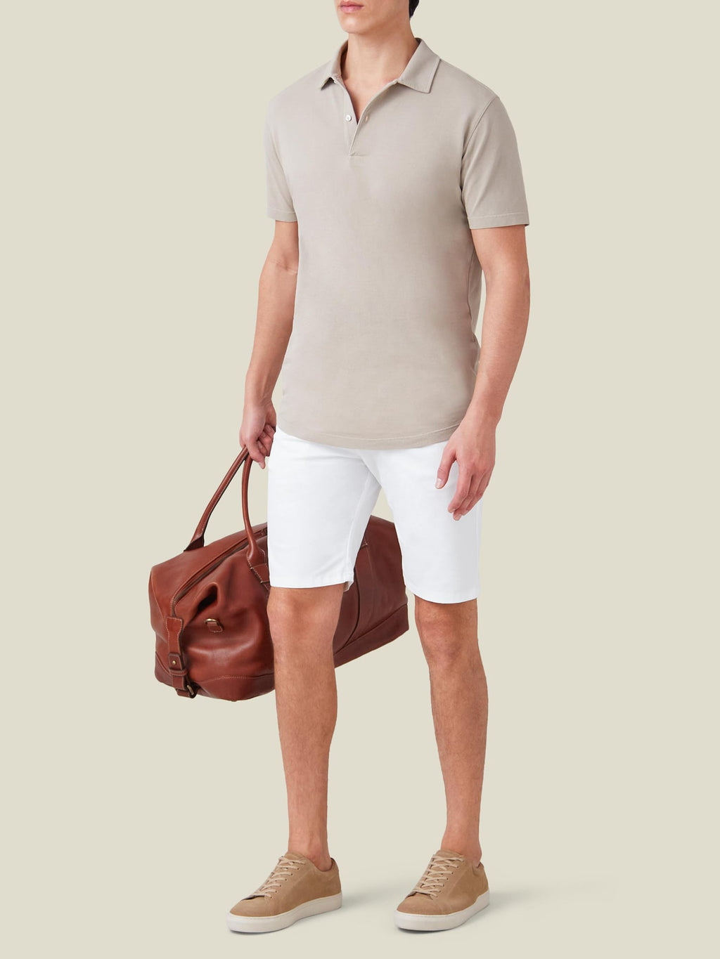 A model wears the Luca Faloni Classic Piqué Polo in Oatmeal with white shorts, sneakers, and a leather travel bag.