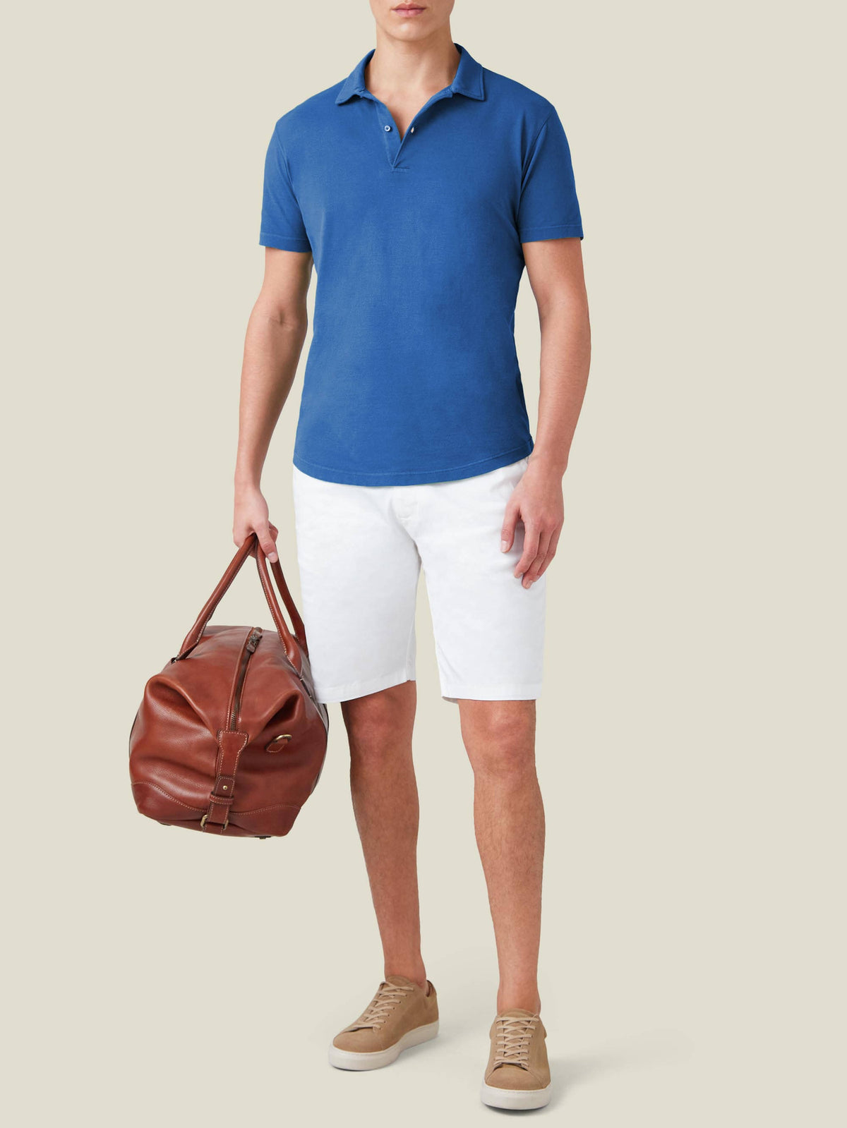 A model wears the Luca Faloni Royal Blue Piqué Polo with white shorts, beige trainers, and a brown leather weekend bag.