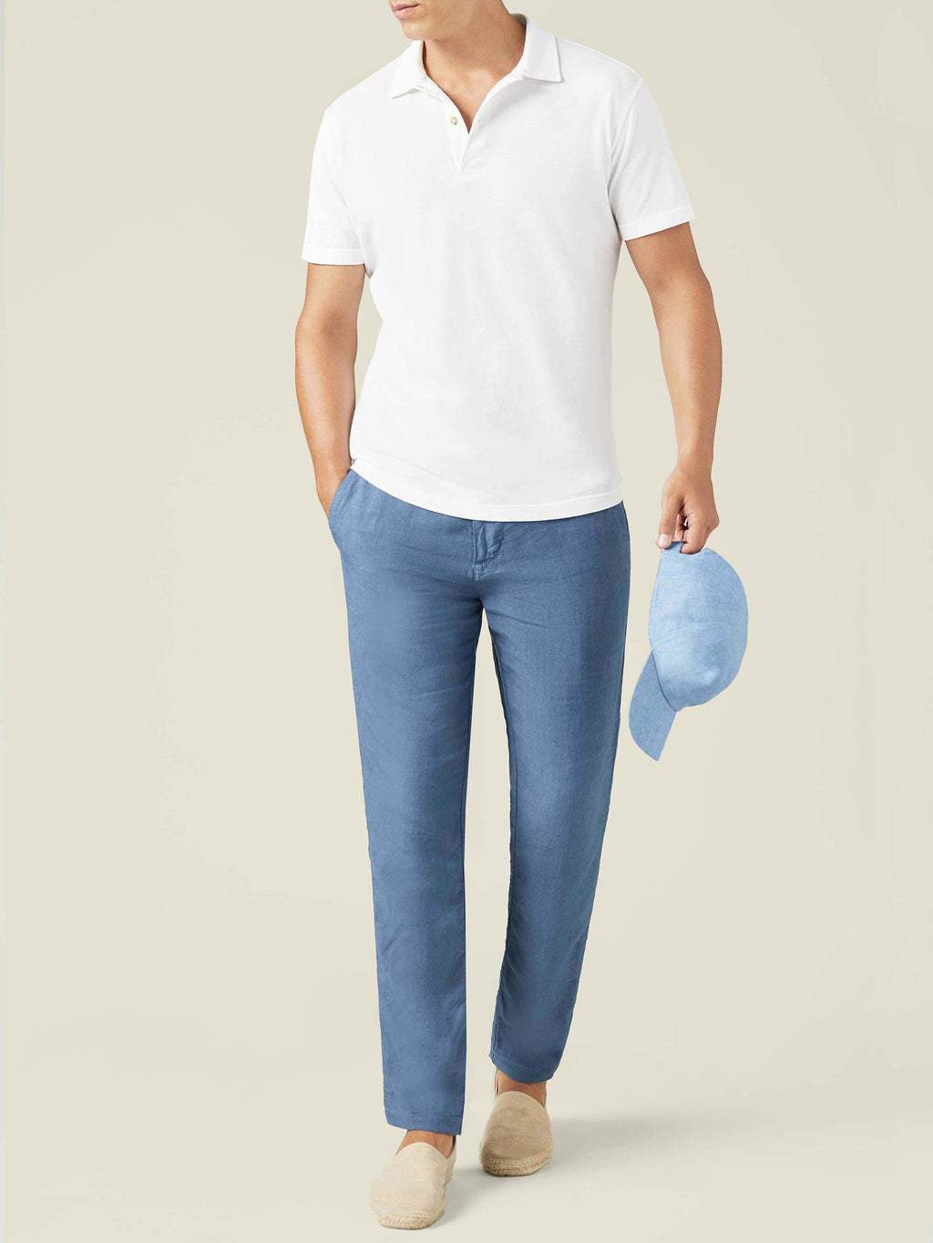 A front view of a model wearing a white piqué polo, blue trousers, and beige espadrilles while holding a light blue cap.