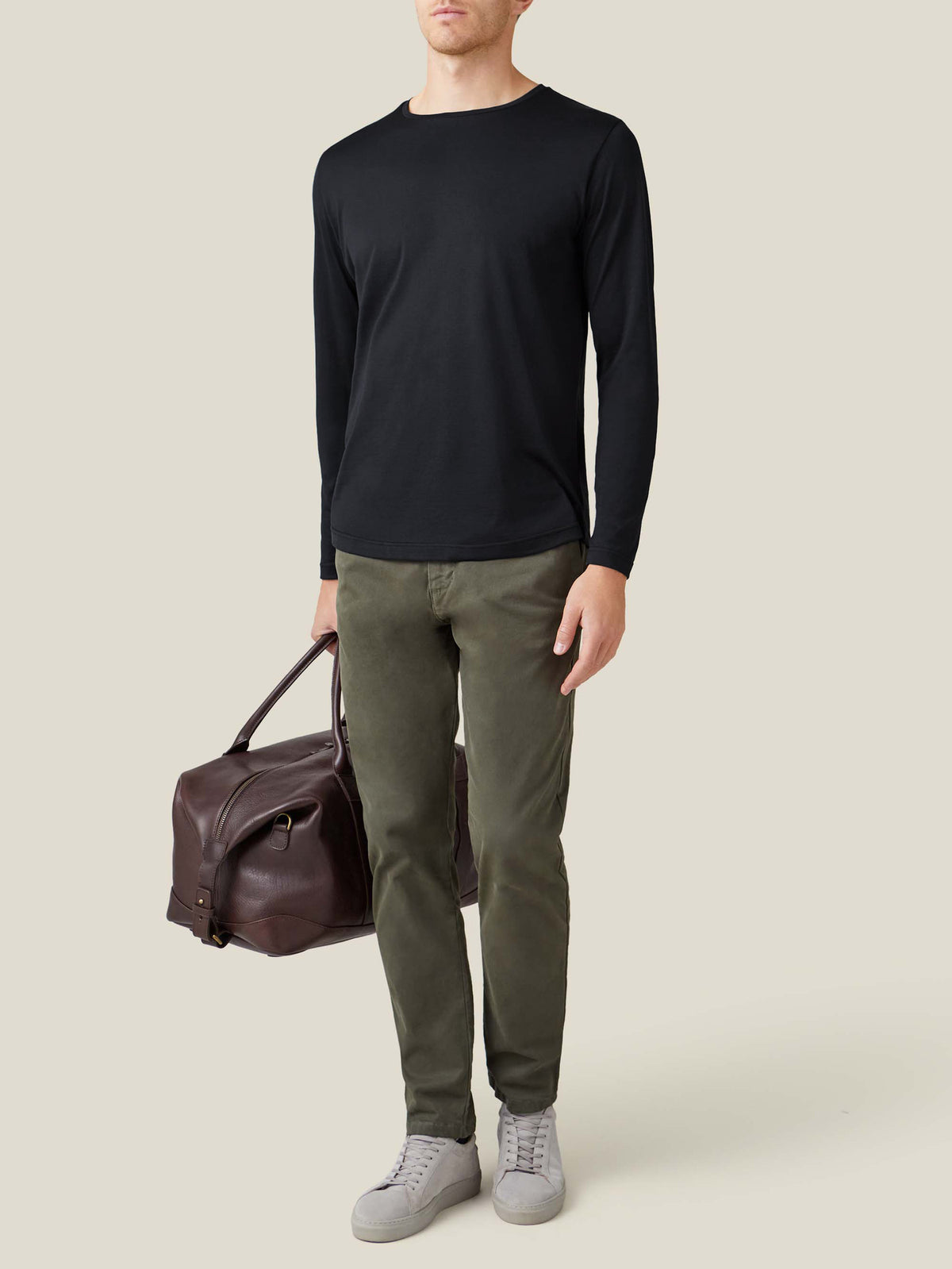 A man wears the black silk-cotton long-sleeved t-shirt, styled with olive chinos, grey trainers, and a brown leather holdall.