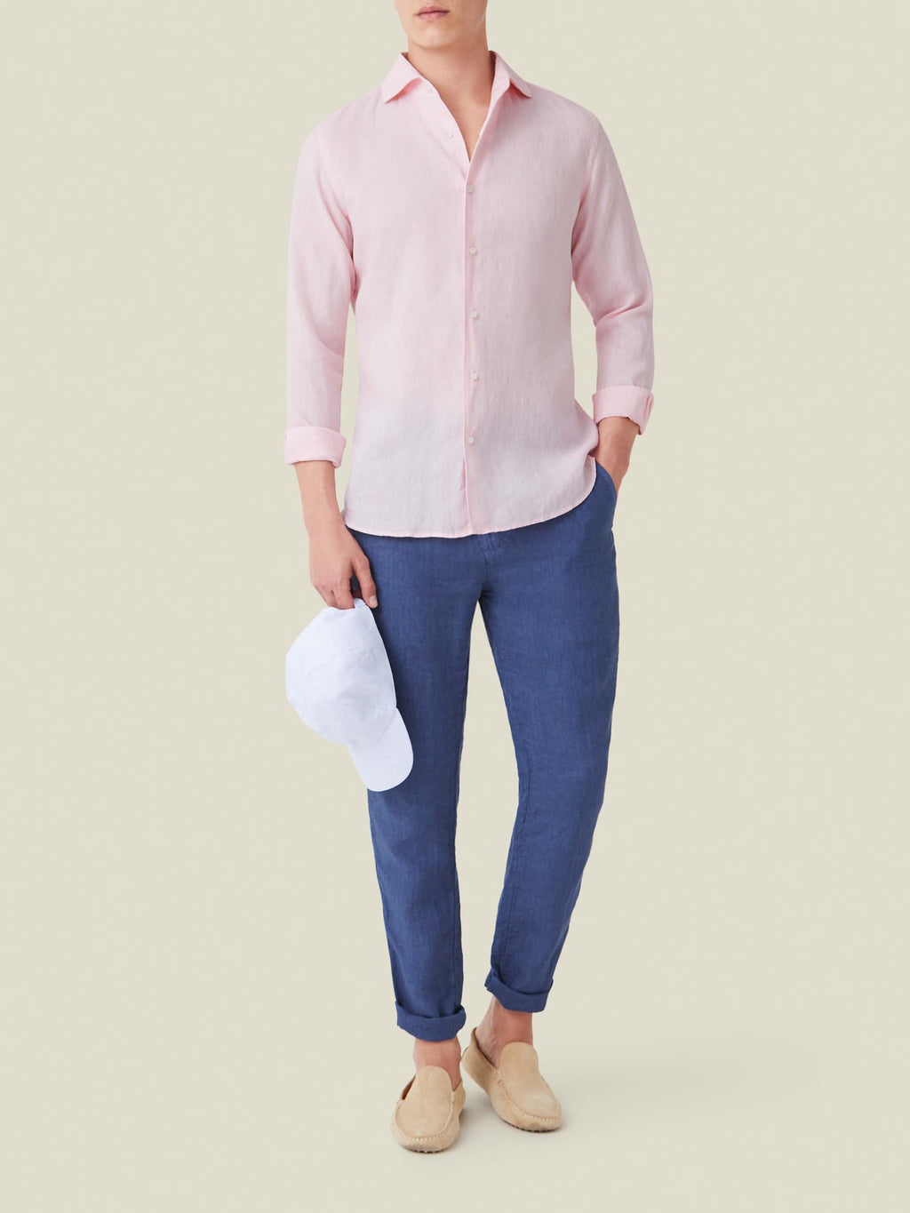 A model wears the Portofino Linen Shirt in Light Pink, styled with blue trousers and beige loafers, holding a white cap.