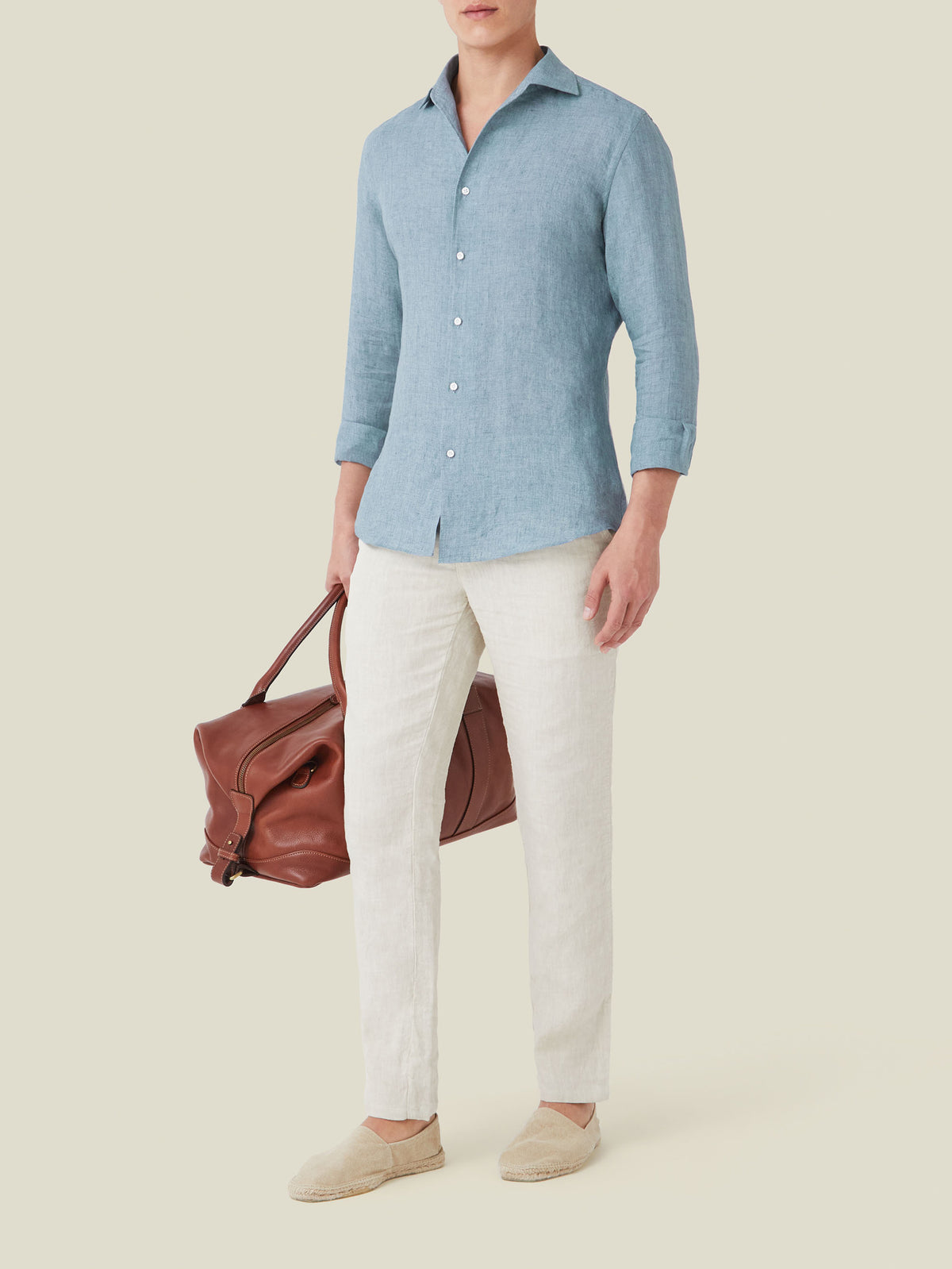 Man models the French Blue Portofino Linen Shirt, styled with white linen trousers, espadrilles, and a brown leather bag.